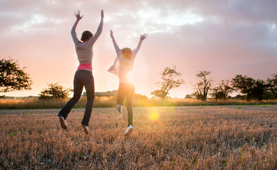 Two girls jumping for joy in sunlit field, rear view - CUF50059