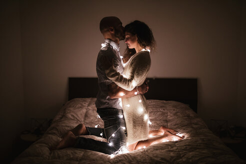 Romantic young couple entwined in decorative lights kneeling face to face on bed - CUF50077