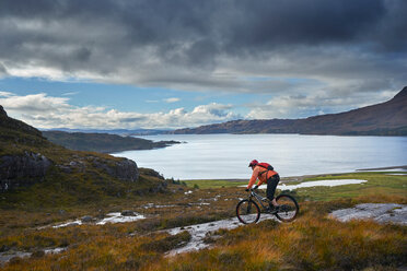 Männlicher Mountainbiker auf dem Fahrrad hinunter zu einem See in einer Berglandschaft, Achnasheen, Schottische Highlands, Schottland - CUF50087