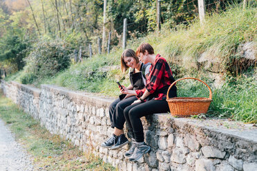Best friends sitting on stone wall, sharing text message, Rezzago, Lombardy, Italy - CUF50095