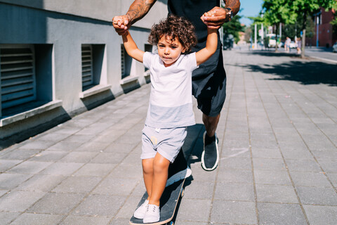 Vater bringt seinem Sohn das Skateboardfahren auf dem Bürgersteig bei, lizenzfreies Stockfoto