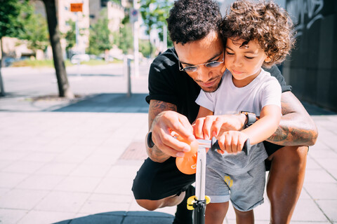 Vater bringt seinem Sohn das Rollerfahren auf dem Bürgersteig bei, lizenzfreies Stockfoto