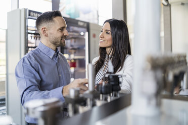 Smiling businessman and businesswoman talking at a machine in modern factory - DIGF06567