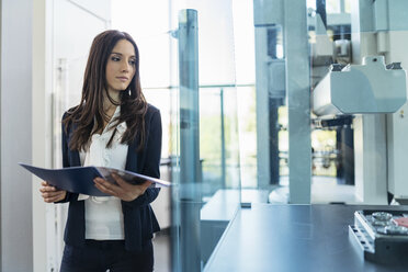 Businesswoman holding folder looking at a machine in modern factory - DIGF06587