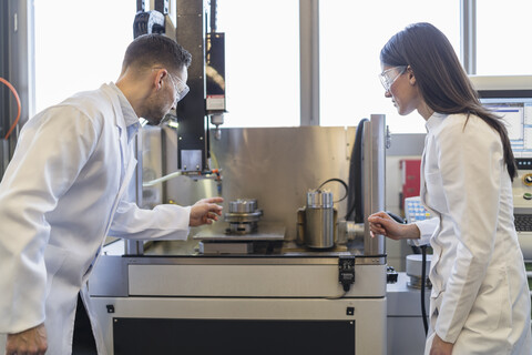 Kollegen in Laborkitteln und mit Schutzbrillen betrachten eine Maschine in einer modernen Fabrik, lizenzfreies Stockfoto