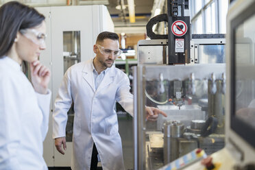 Colleagues wearing lab coats and safety goggles looking at machine in modern factory - DIGF06711