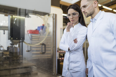 Colleagues wearing lab coats and safety goggles looking at machine in modern factory - DIGF06714