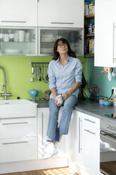 Reife Frau mit einer Tasse Kaffee auf dem Küchentisch sitzend zu Hause - FLLF00089