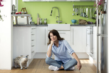 Portrait of mature woman sitting on the floor in kitchen at home - FLLF00096