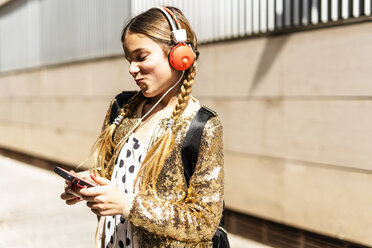 Lächelndes Mädchen mit goldener Paillettenjacke und Kopfhörern, das auf sein Handy schaut - ERRF00899