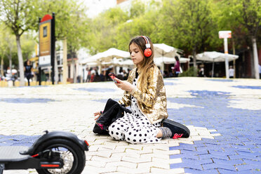 Mädchen hockt auf einem Platz und hört Musik mit Kopfhörern und Smartphone - ERRF00932
