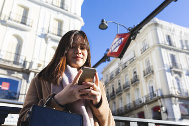Spain, Madrid, smiling young woman at Puerta del Sol metro station checking her smartphone - WPEF01463