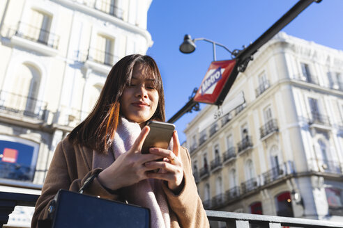 Spanien, Madrid, lächelnde junge Frau an der U-Bahn-Station Puerta del Sol, die ihr Smartphone überprüft - WPEF01463