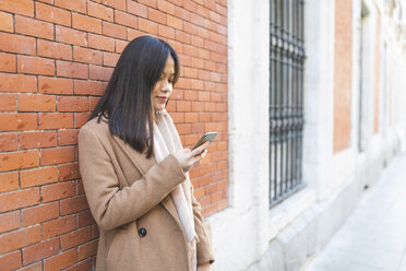 Young woman using cell phone at brick wall - WPEF01472