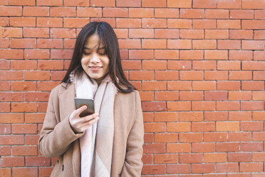 Lächelnde junge Frau, die ein Handy an einer Backsteinmauer benutzt - WPEF01473