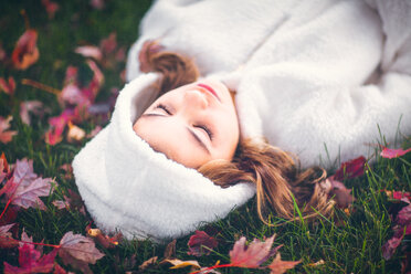 Girl in white hooded top with eyes closed, lying on grass amongst autumn leaves, portrait - ISF21162