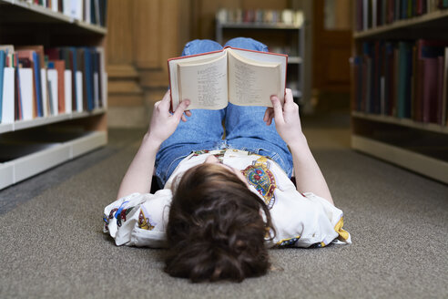 Female student reading book in a public library, lying on the ground - IGGF01047