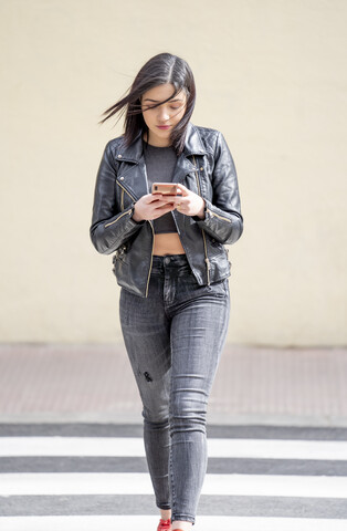 Junge Frau, die den Fußgängerüberweg überquert und ein Mobiltelefon benutzt, lizenzfreies Stockfoto