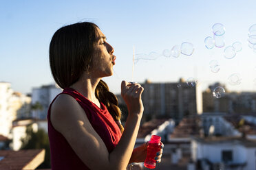 Teenage girl blowing soap bubbles on roof terrace in the city at sunset - ERRF00938
