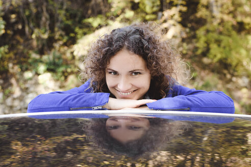 Porträt einer lächelnden Frau mit lockigem Haar, die sich auf ein Autodach stützt - EYAF00140