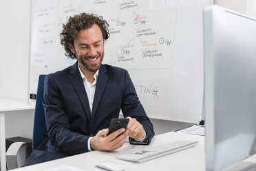 Smiling businessman sitting at desk in office using cell phone - MGIF00374