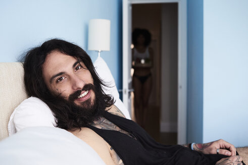 Porträt eines lächelnden, tätowierten Mannes, der im Bett liegt, mit einer Freundin im Hintergrund - IGGF01095