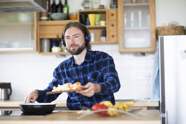 Junger Mann mit Bart und kariertem Hemd und Kopfhörer beim Kochen von Gemüse in der Küche - SGF02362