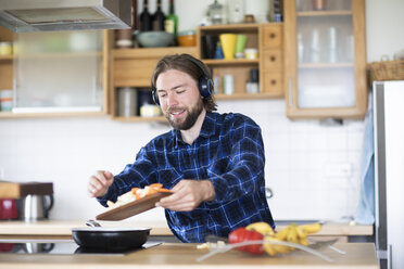 Junger Mann mit Bart und kariertem Hemd und Kopfhörer beim Kochen von Gemüse in der Küche - SGF02363