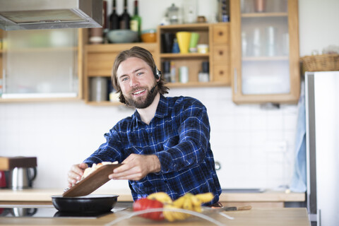 Junger Mann mit Bart, kariertem Hemd und Headset, der in der Küche Gemüse kocht, lizenzfreies Stockfoto