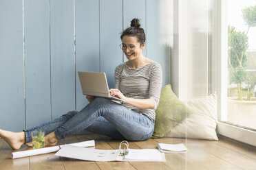 Smiling woman sitting at the window at home working with laptop and file folder - UUF17165