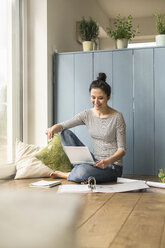 Smiling woman sitting at the window at home working with laptop and file folder - UUF17169