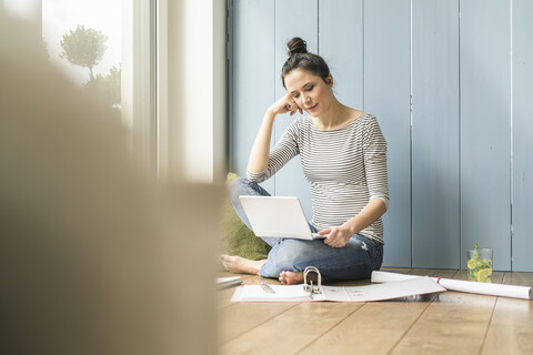 Frau sitzt zu Hause am Fenster und arbeitet mit Laptop und Aktenordner, lizenzfreies Stockfoto