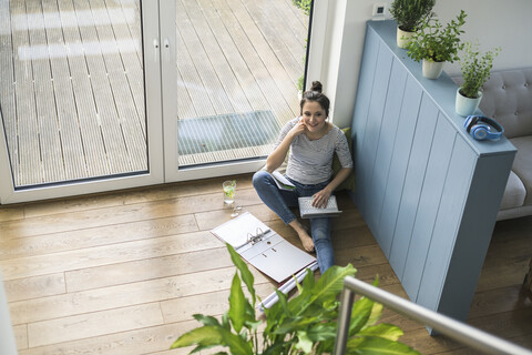 Bird's eye view of smiling woman sitting at the window at home working with laptop and file folder stock photo