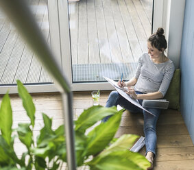 Bird's eye view of smiling woman sitting at the window at home working with laptop and file folder - UUF17196