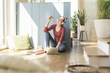 Woman with headphones sitting at the window at home using laptop - UUF17197