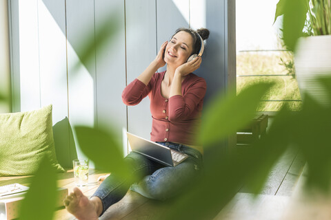 Lächelnde Frau mit Kopfhörern und Laptop, die zu Hause am Fenster sitzt, lizenzfreies Stockfoto