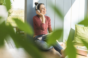 Happy woman with headphones and laptop sitting at the window at home - UUF17203