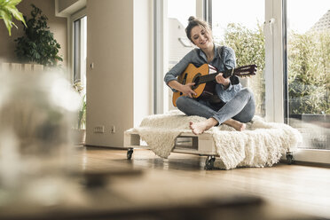 Smiling woman sitting at the window at home playing guitar - UUF17204
