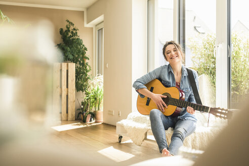 Glückliche Frau, die zu Hause am Fenster sitzt und Gitarre spielt - UUF17205