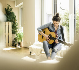Woman sitting at the window at home playing guitar - UUF17206