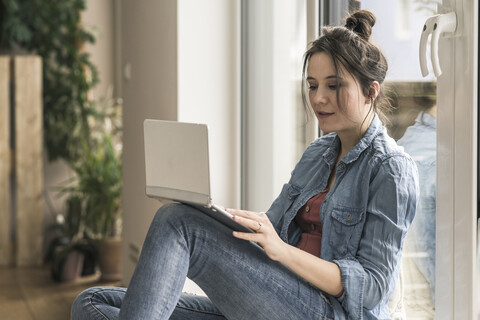 Frau sitzt zu Hause am Fenster und benutzt einen Laptop, lizenzfreies Stockfoto