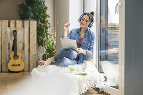 Frau mit Kopfhörern und Laptop sitzt zu Hause am Fenster, lizenzfreies Stockfoto