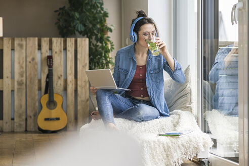 Frau mit Kopfhörern und Laptop sitzt zu Hause am Fenster und trinkt aus einem Glas - UUF17211