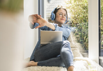 Woman with headphones and laptop sitting at the window at home - UUF17212