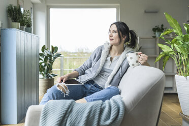 Nachdenkliche Frau mit Tasse und Tablet auf der Couch zu Hause sitzend - UUF17216