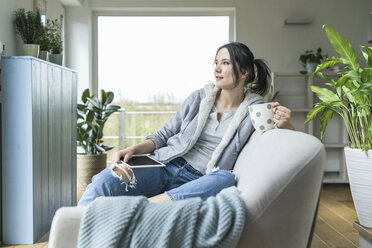 Pensive woman with a mug and tablet sitting on the couch at home - UUF17216