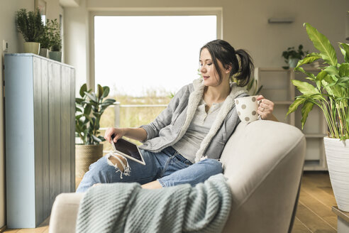 Frau mit einem Becher sitzt auf der Couch zu Hause mit Tablet - UUF17218
