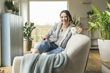 Porträt einer glücklichen Frau mit einer Tasse und einem Tablet, die zu Hause auf der Couch sitzt - UUF17219
