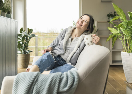 Glückliche Frau mit einer Tasse und einem Tablet, die zu Hause auf der Couch sitzt - UUF17220