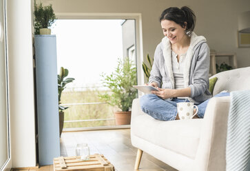 Woman with a mug sitting on the couch at home using tablet - UUF17221